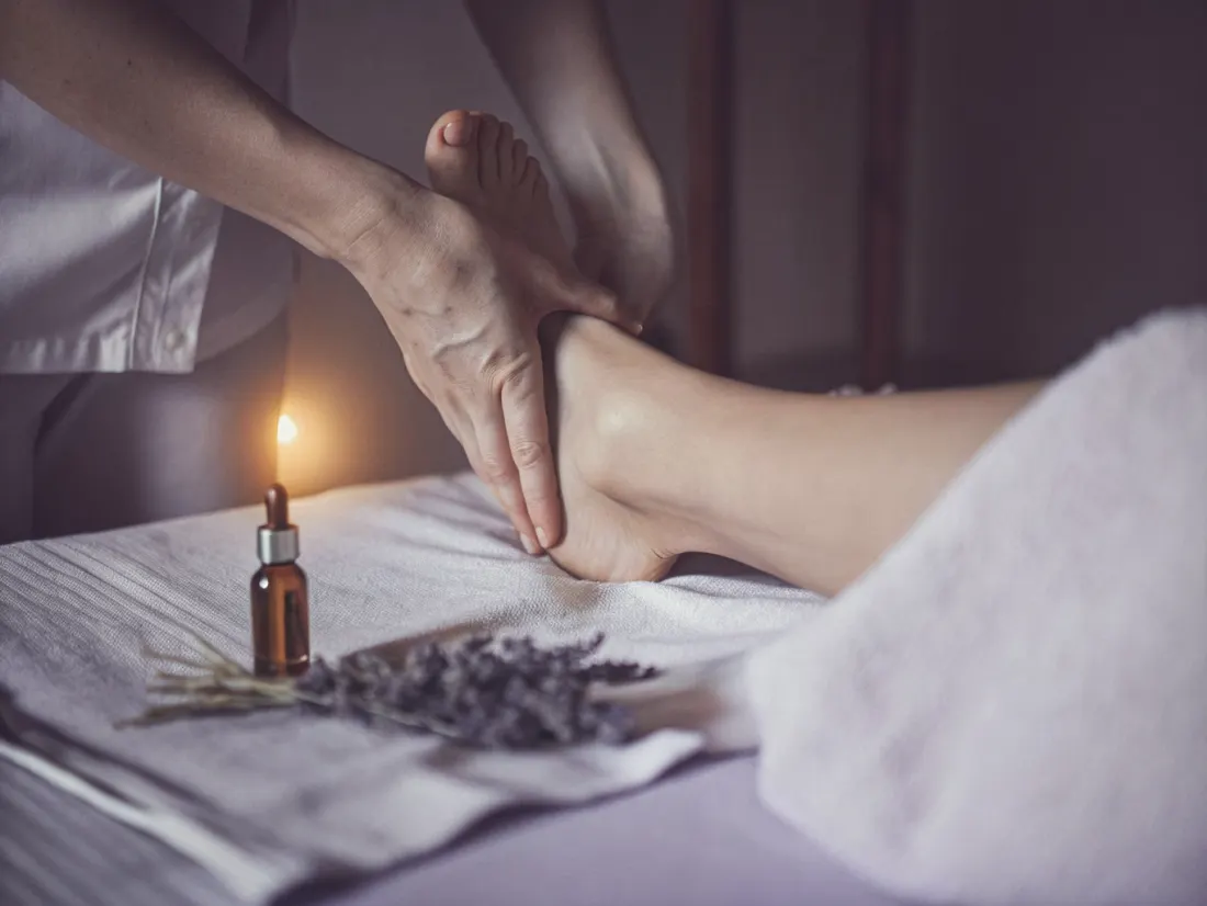 Mãos de uma terapeuta a massajar o pé de uma cliente, com um frasco de óleo essencial e um ramo de lavanda